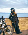 Person in motorcycle gear standing next to a motorcycle on a coastal road.