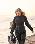 A blond young woman walking on a beach and carrying a motorcycle helmet wearing Black and grey lumberjack style women's motorcycle shirt from Shima
