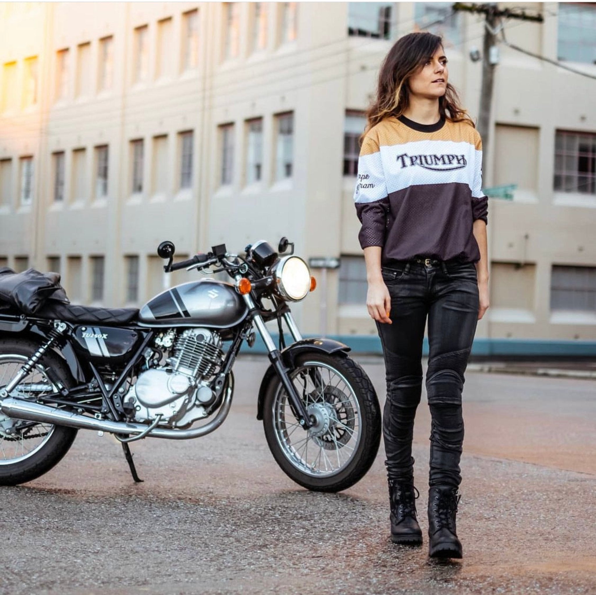 A woman wearing black leather boots and Triumph shirt walking away from her motorcycle