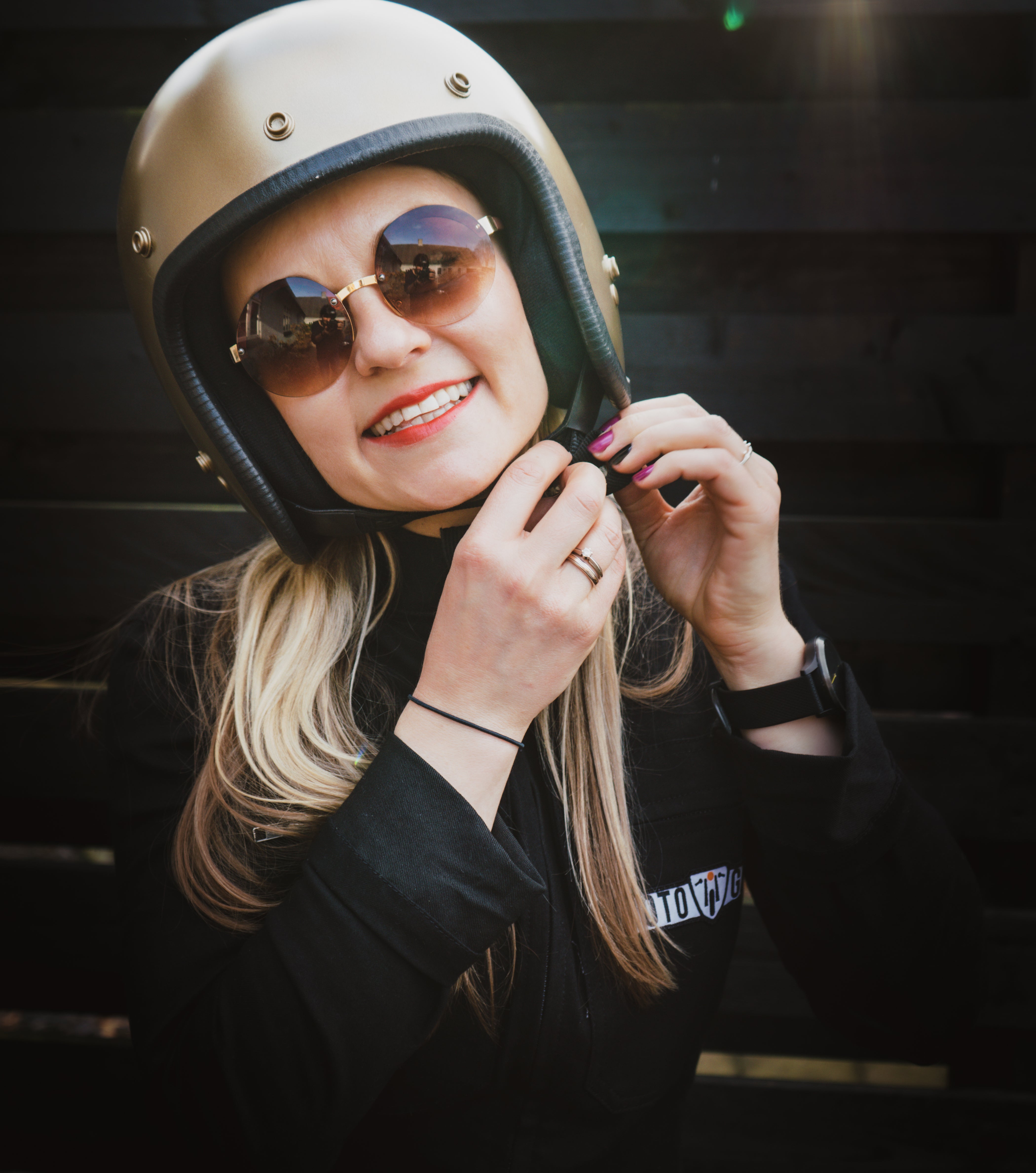 A smiling women wearing round sunglasses and Moto Girl overall putting golden motorcycle helmet on
