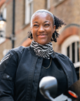 A woman wearing a black motorcycle jacket and a black-and-white patterned neck gaiter with the word 'Motogadget' printed on it, standing outdoors in front of a brick building.