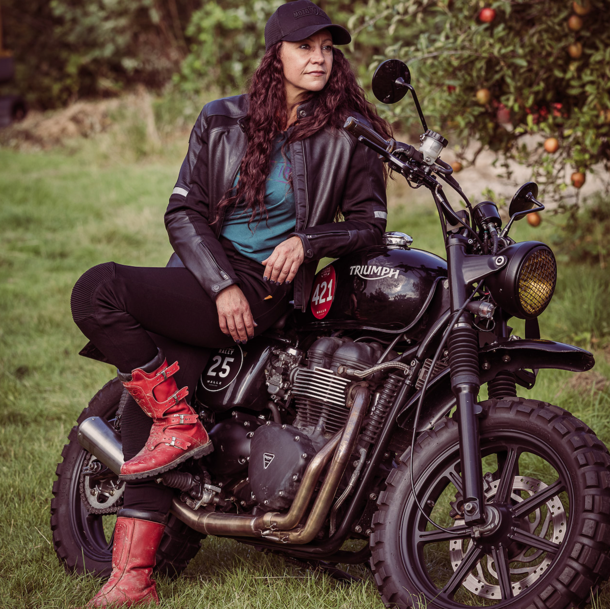 A woman leaning on her Triumph motorcycle wearing black leather motorcycle jacket and red boots