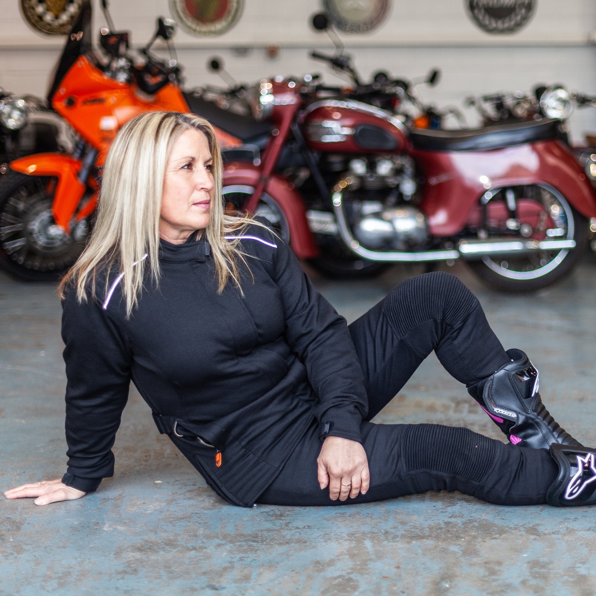 A blong woman sitting on the floor by motorcycles wearing women's motorcycle jacket from Moto Girl