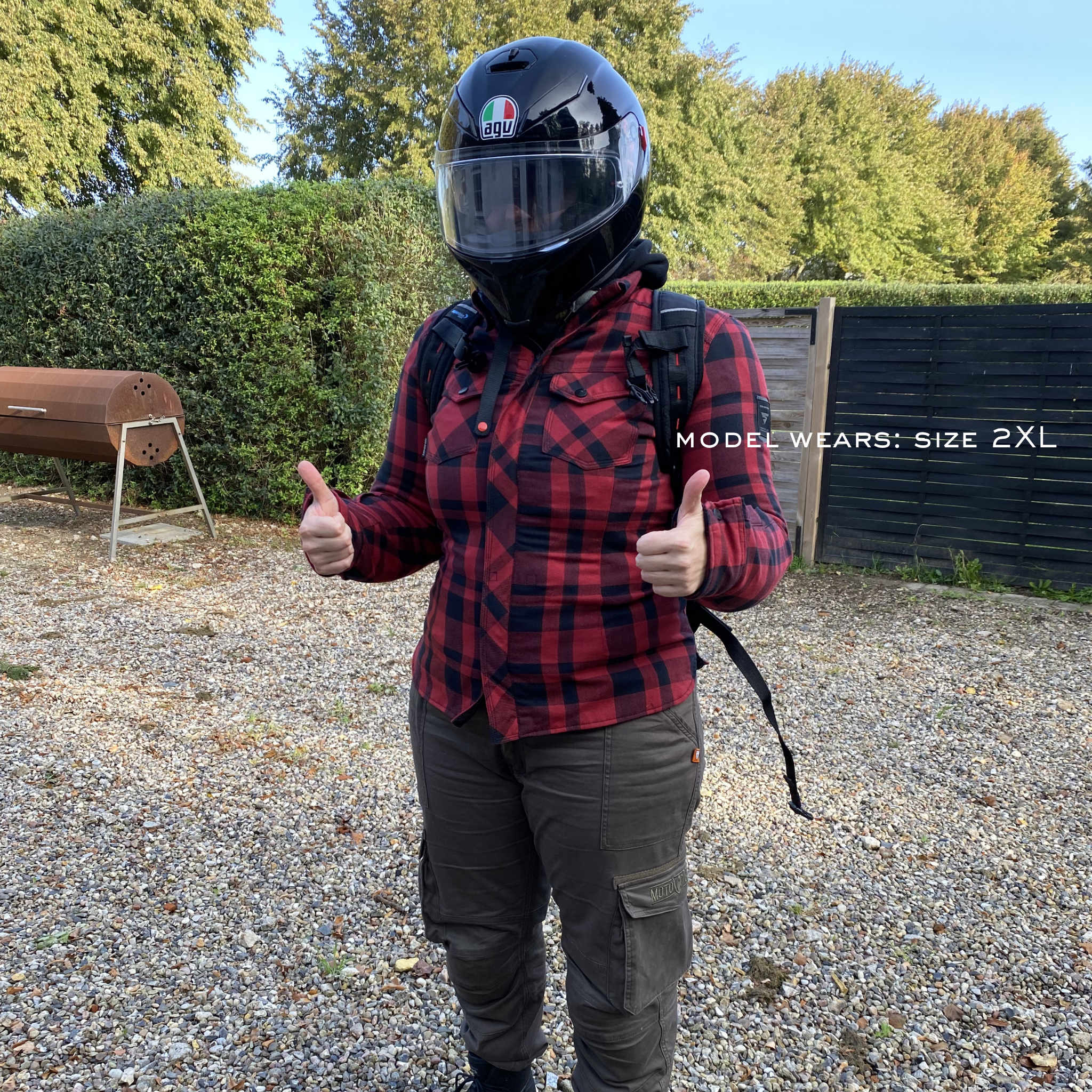Person wearing a red plaid shirt, black helmet, and backpack giving a thumbs-up gesture outdoors.