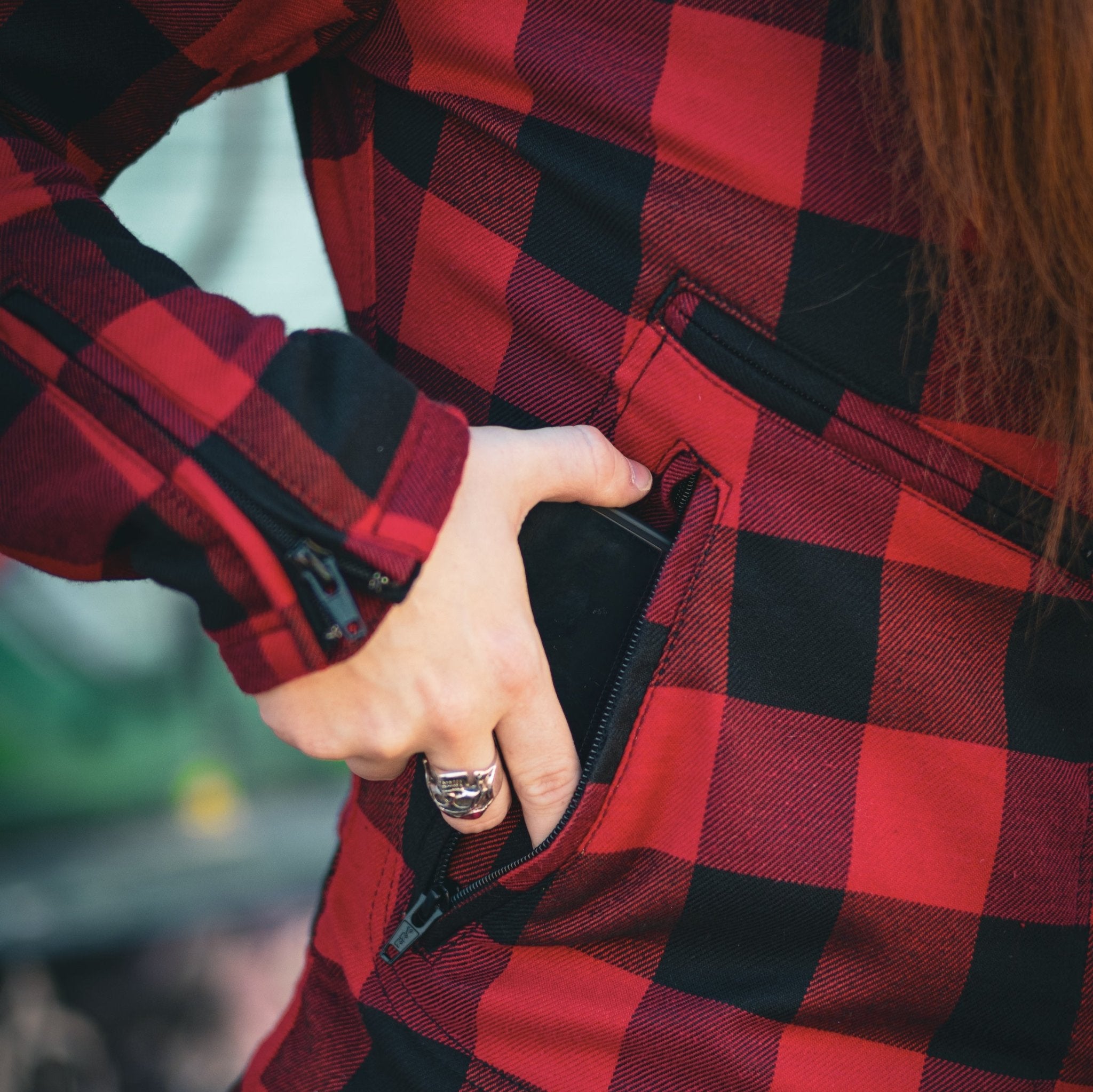 Close-up of a woman wearing a red and black flannel-style women's jacket, putting a phone in the side pocket.