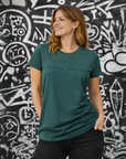 Woman wearing a green 'Moto Girl' t-shirt standing in front of a graffiti-covered wall.