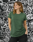 Woman wearing a green t-shirt with 'MotoGirl' logo against a graffiti-covered wall.