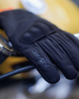 BLACK MOTORCYCLE GLOVES WITH MOTOGIRL LOGO ON A HANDLE BAR OF A MOTORCYCLE