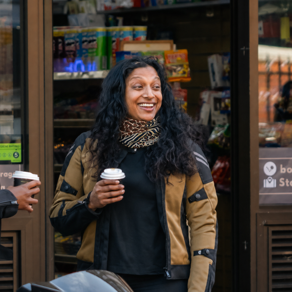 A woman smiling and holding a takeaway coffee cup stands in front of a convenience store entrance. She is wearing a tan and black motorcycle-style jacket over a black top and a patterned scarf.