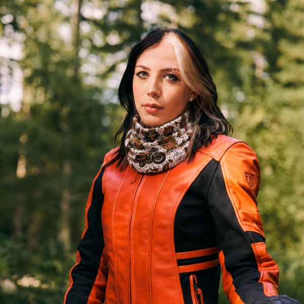 A woman wearing a bright orange and black leather motorcycle jacket and a patterned knit neck warmer, standing outdoors in a blurred green forest setting.