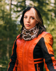 A woman wearing a bright orange and black leather motorcycle jacket and a patterned knit neck warmer, standing outdoors in a blurred green forest setting.