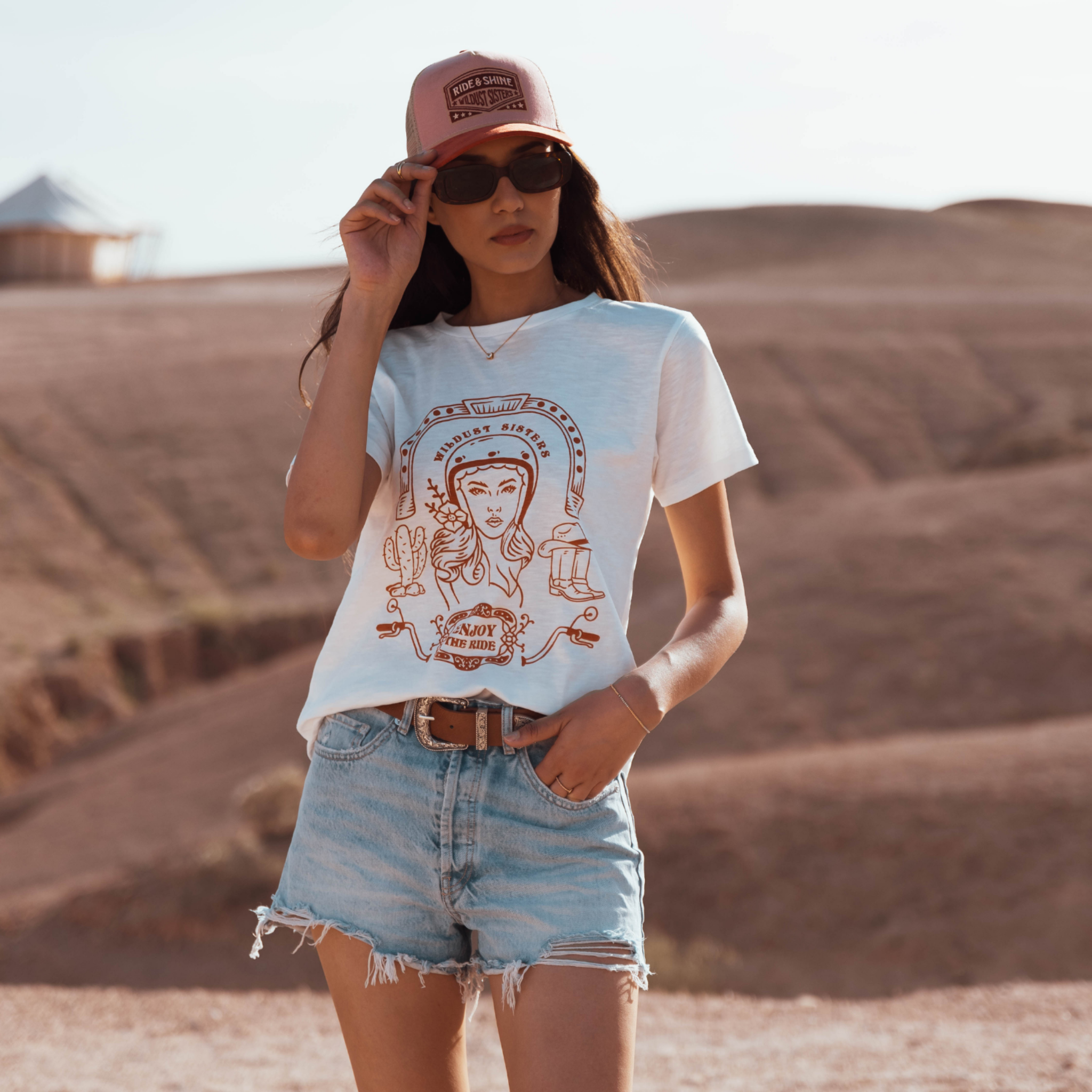 Woman in the dessert wearing a white T-shirt with a Woman wearing a Motorcyclehelmet print and the text &quot;Wild dust sister&quot; and &quot;Enjoy The ride&quot; in orange