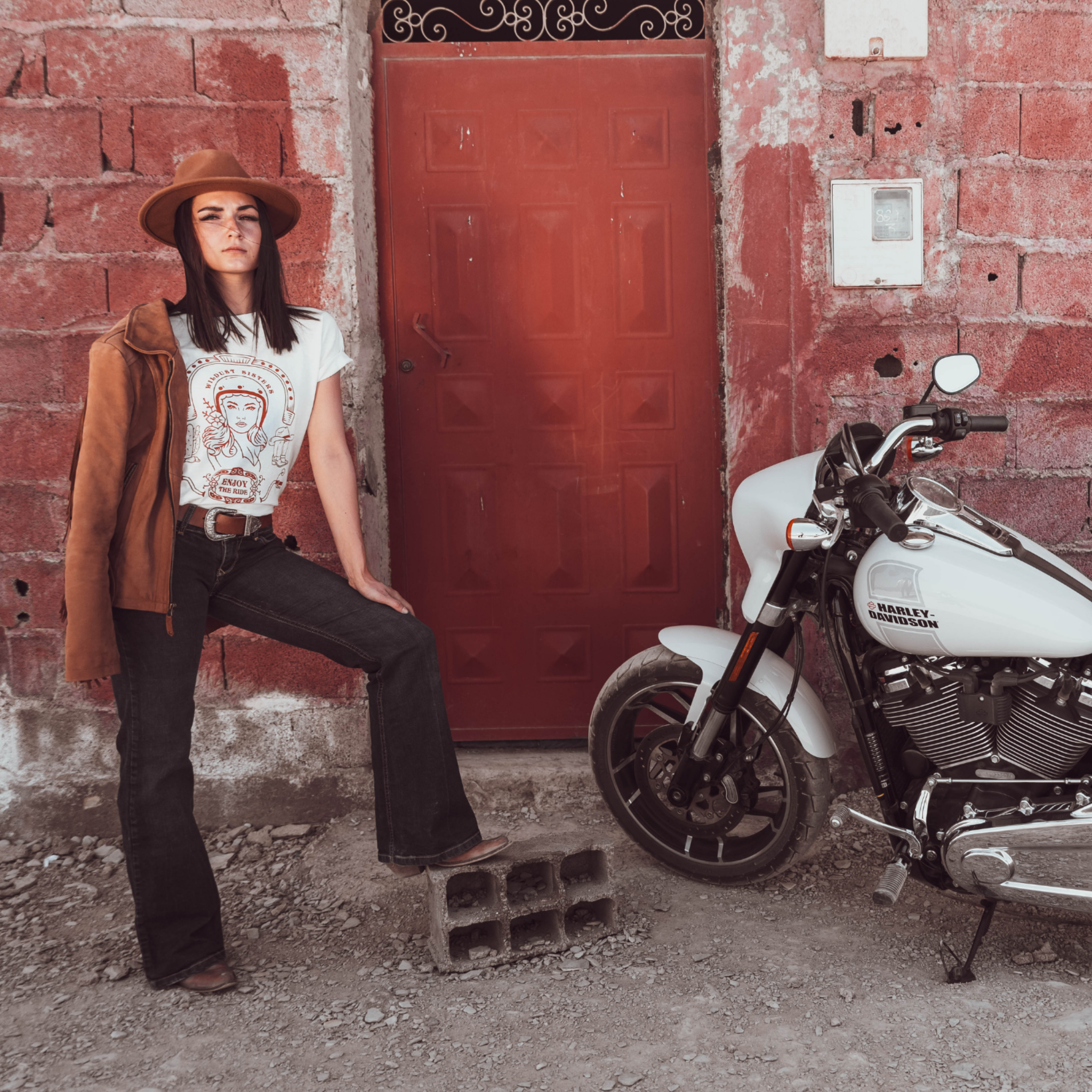 Woman standing next to a Motorcycle wearing a white T-shirt with a Woman wearing a Motorcyclehelmet print and the text "Wild dust sister" and "Enjoy The ride" in orange