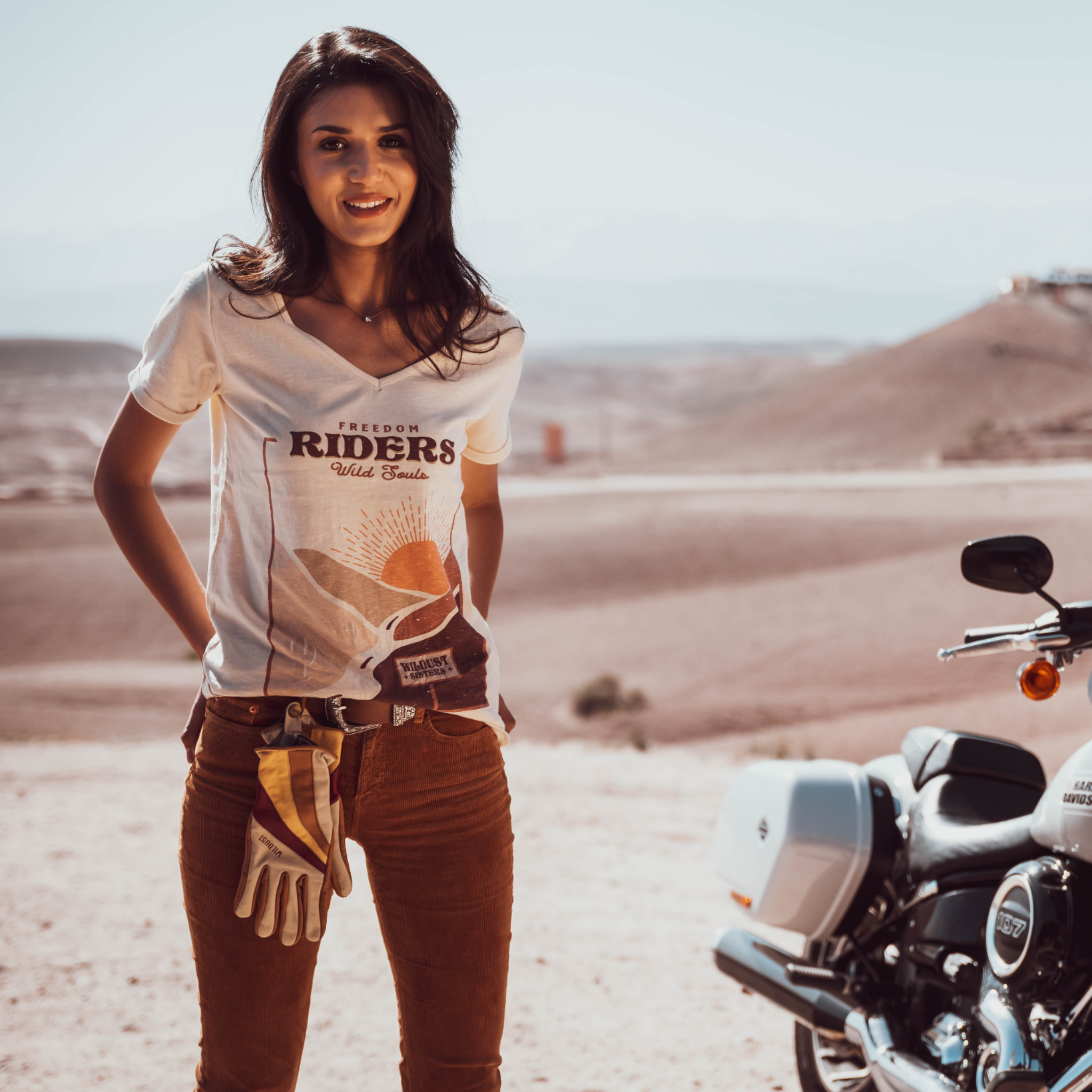 Woman standing in the dessert next to a Motorcycle wearing a T-shirt with a sunset/sunrise print and the text &quot;Freedom Riders Wild Souls&quot; and &quot;Wildust sisters&quot;