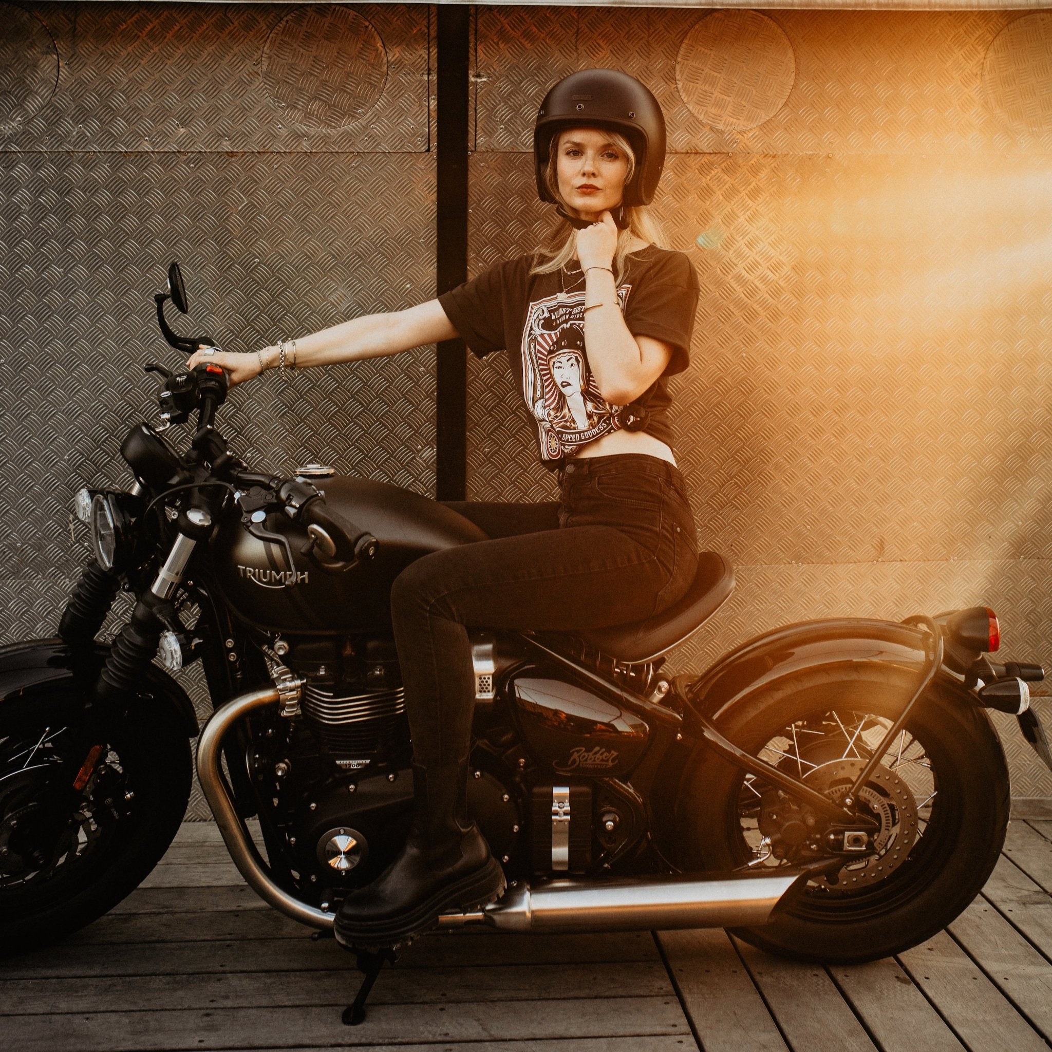 Woman sitting on a Motorcycle wearing  a T-shirt with a women wearing a Motorcyclehelmet print and with the text &quot;Wild dust sisters woman rider&quot; and &quot;Speed Goddess&quot;