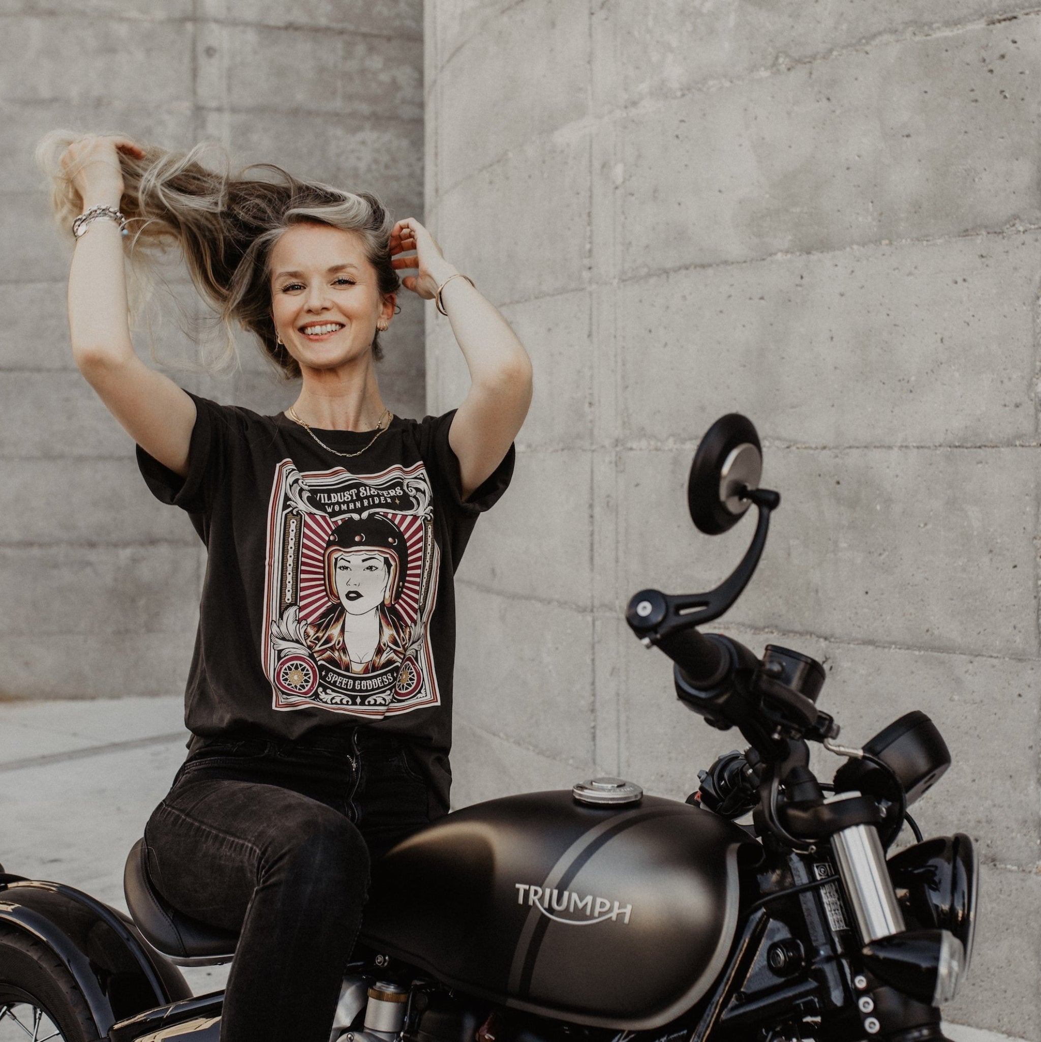 Woman sitting on a Motorcycle wearing  a T-shirt with a women wearing a Motorcyclehelmet print and with the text "Wild dust sisters woman rider" and "Speed Goddess"