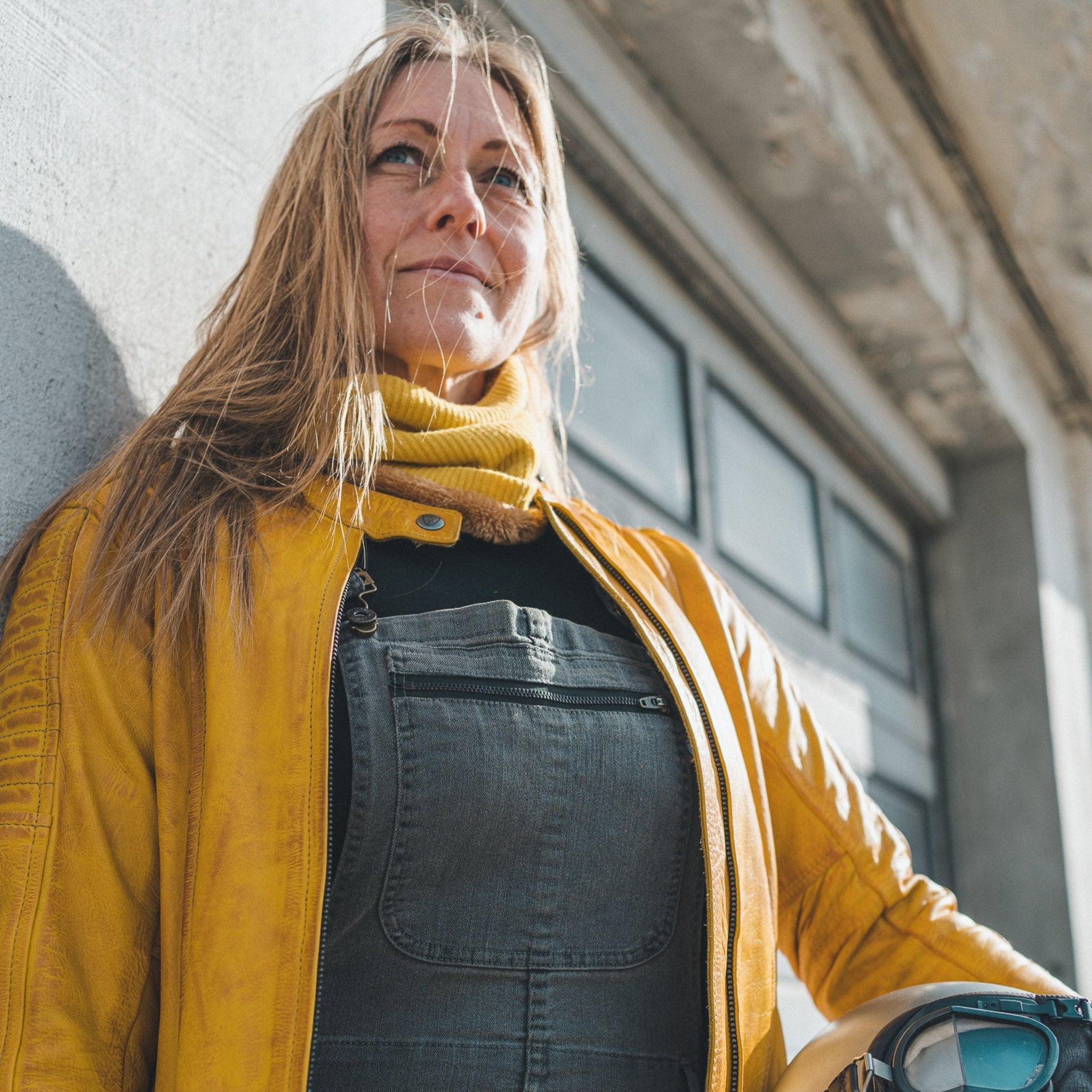 Woman standing outside against a wall, wearing a yellow Valerie jacket and a yellow knitted neck warmer from Moto Girl.