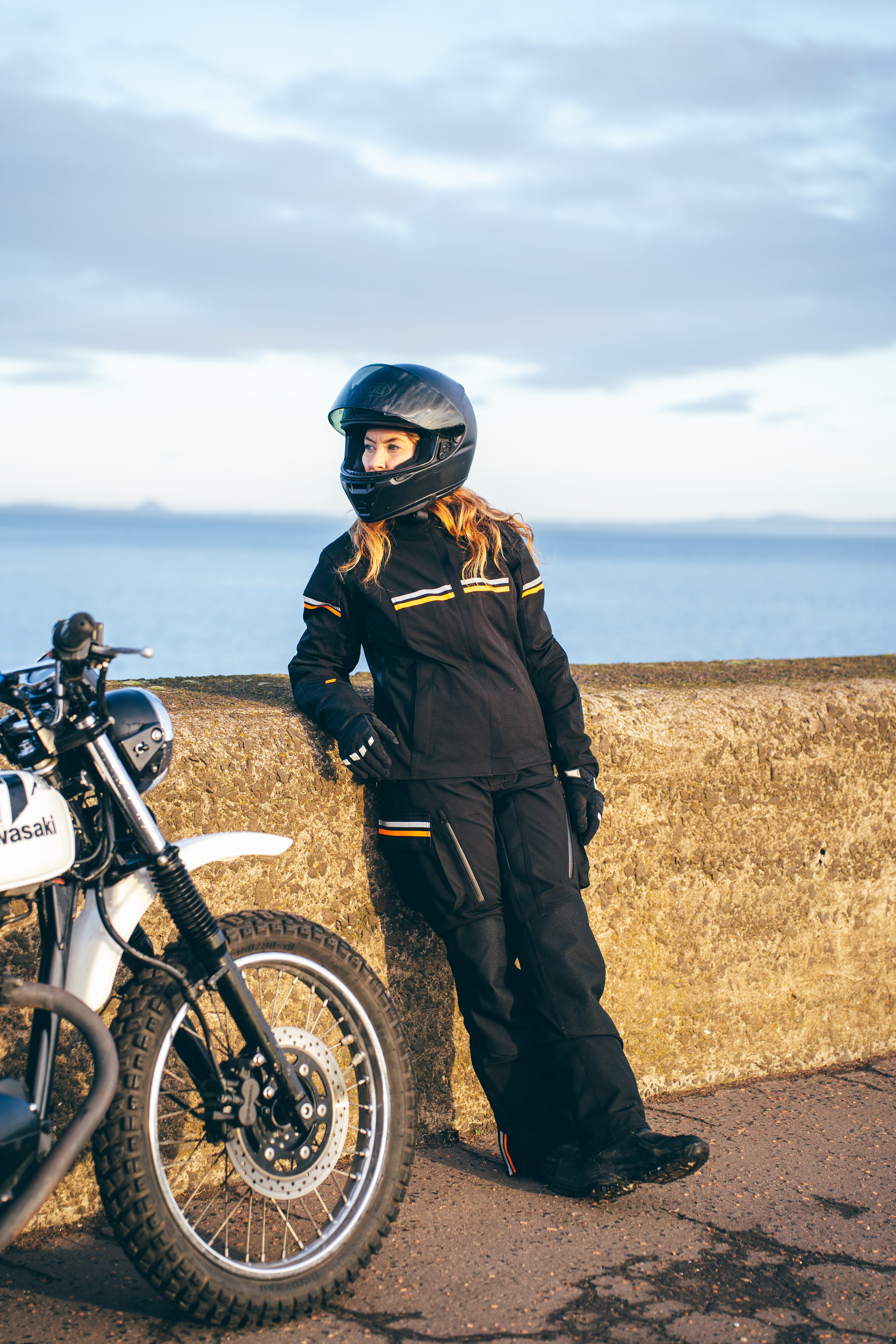 Person in motorcycle gear standing next to a motorcycle on a coastal road.
