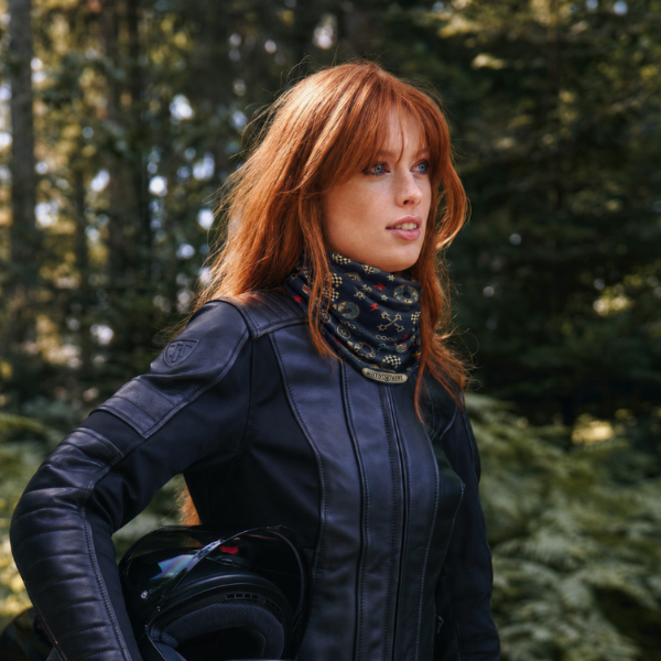 A woman with red hair wearing a black leather motorcycle jacket and a patterned neck gaiter, holding a black helmet in a forest setting.