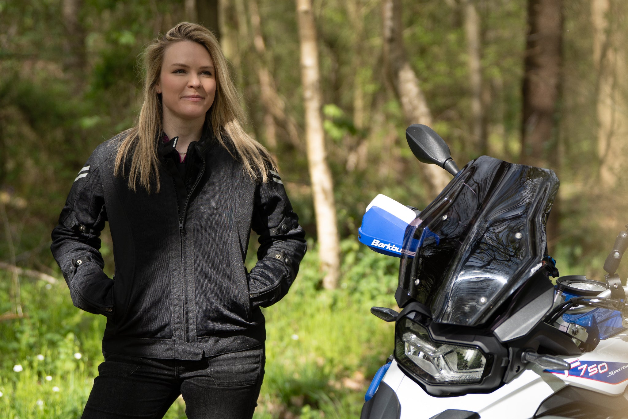 a blong smiling woman standing by her big motorcycle wearing a black mesh motorcycle jacket for women