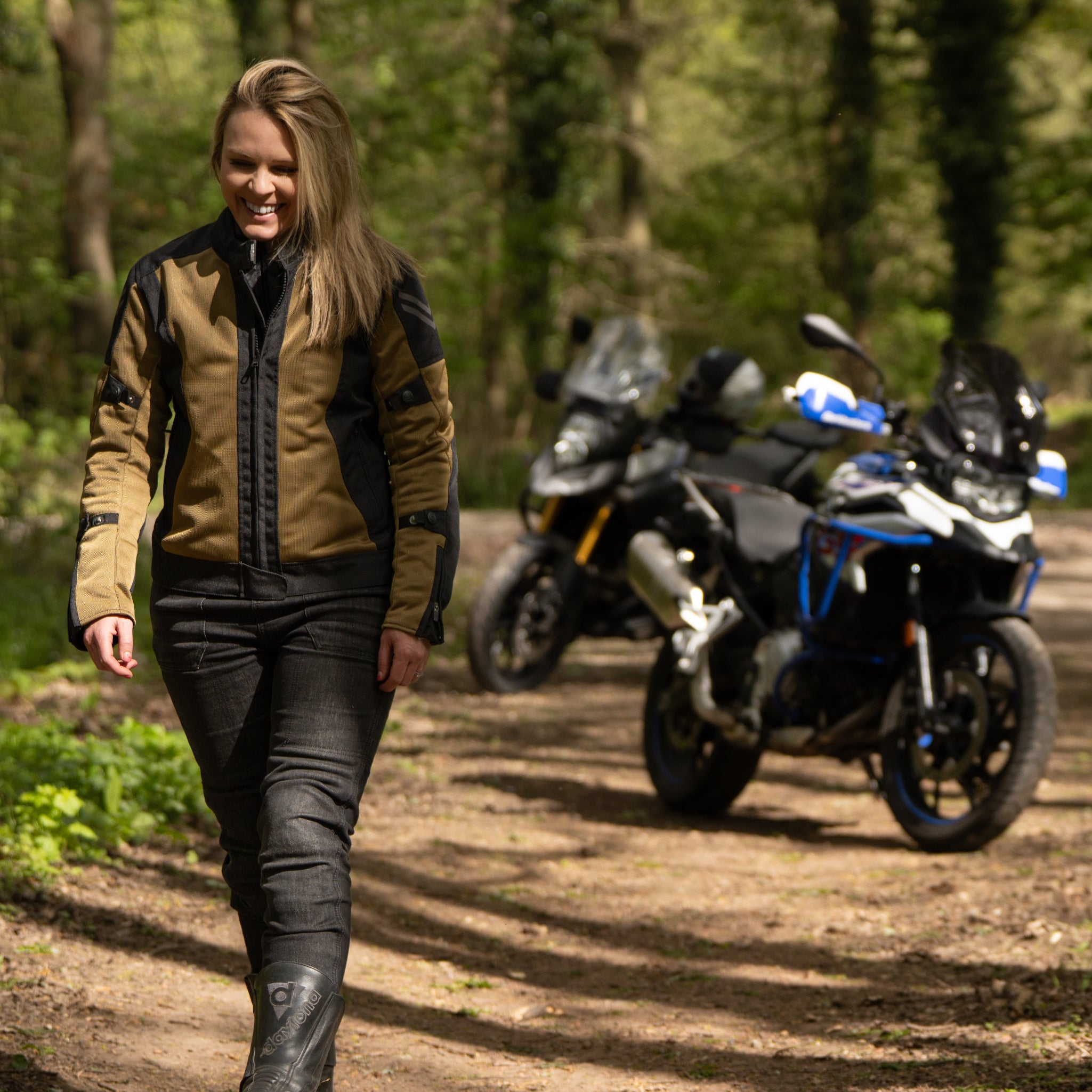 a blond smiling woman walking away from her motorcycle wearing a black and gold mesh motorcycle jacket for women