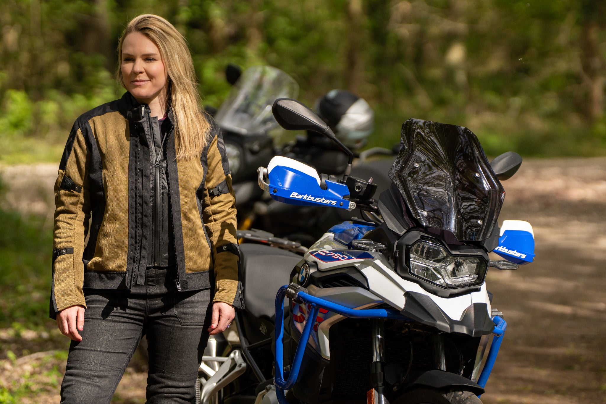 a blond smiling woman standing by her big motorcycle wearing a black and gold mesh motorcycle jacket for women