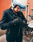 Person wearing a black jacket and helmet, standing next to a motorcycle with a red wall in the background.