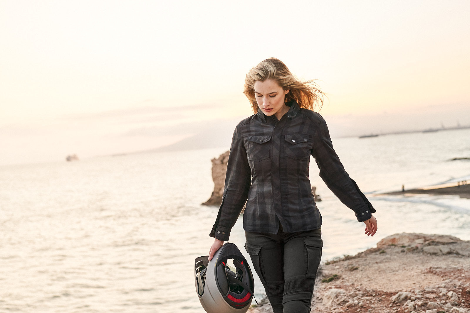 A blond young woman walking on a beach and carrying a motorcycle helmet wearing Black and grey lumberjack style women&#39;s motorcycle shirt from Shima
