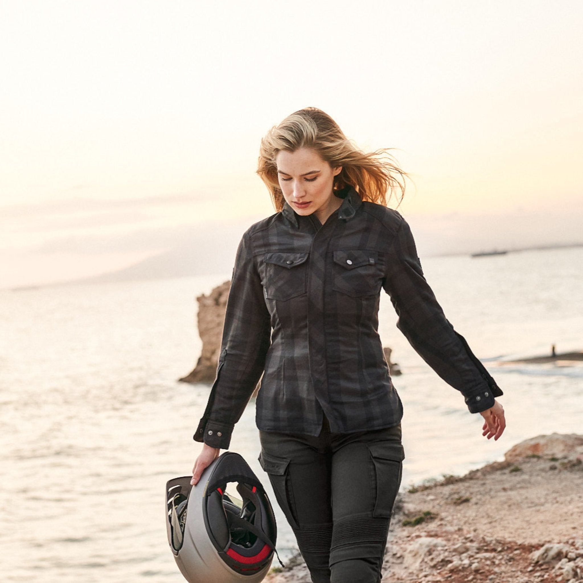 A blond young woman walking on a beach and carrying a motorcycle helmet wearing Black and grey lumberjack style women's motorcycle shirt from Shima