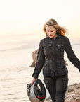 A blond young woman walking on a beach and carrying a motorcycle helmet wearing Black and grey lumberjack style women's motorcycle shirt from Shima