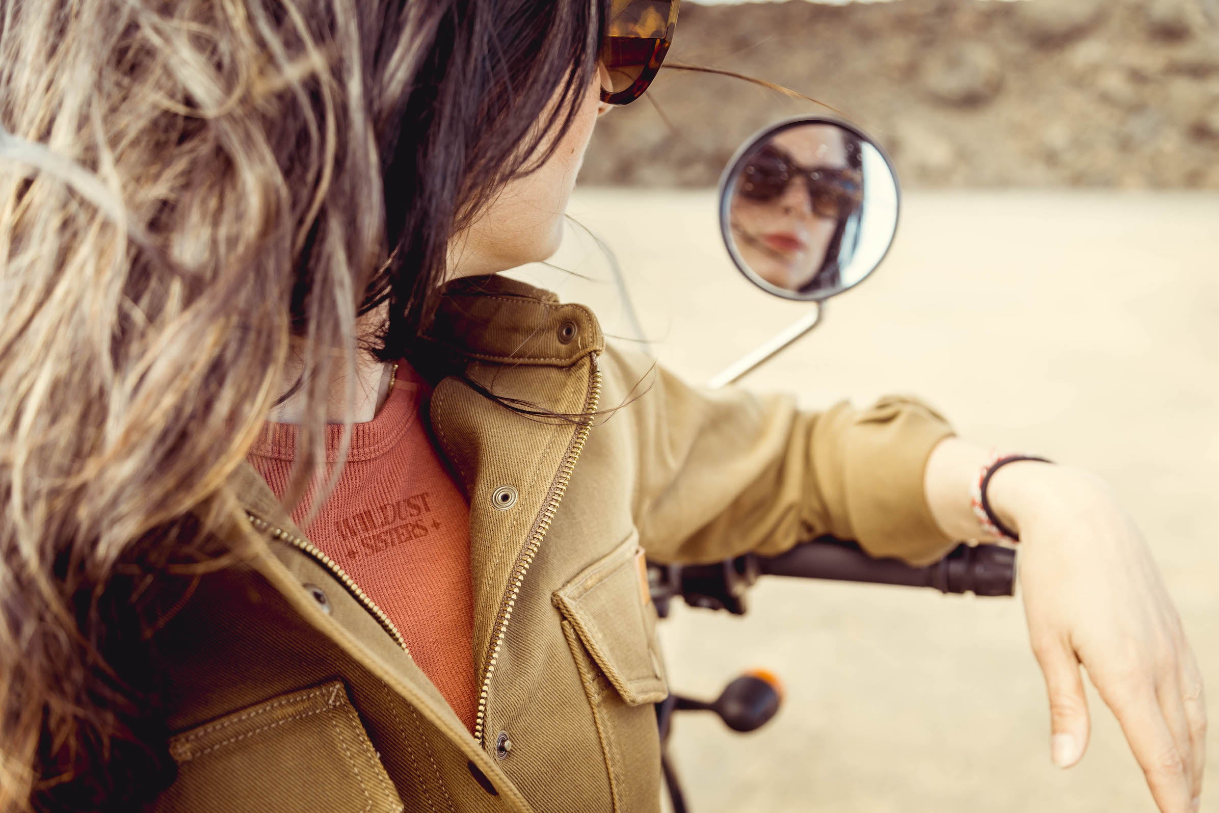 a woman looking to the side mirror on her motorcycle wearing brown khaki denim mc shirt