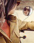 a woman looking to the side mirror on her motorcycle wearing brown khaki denim mc shirt