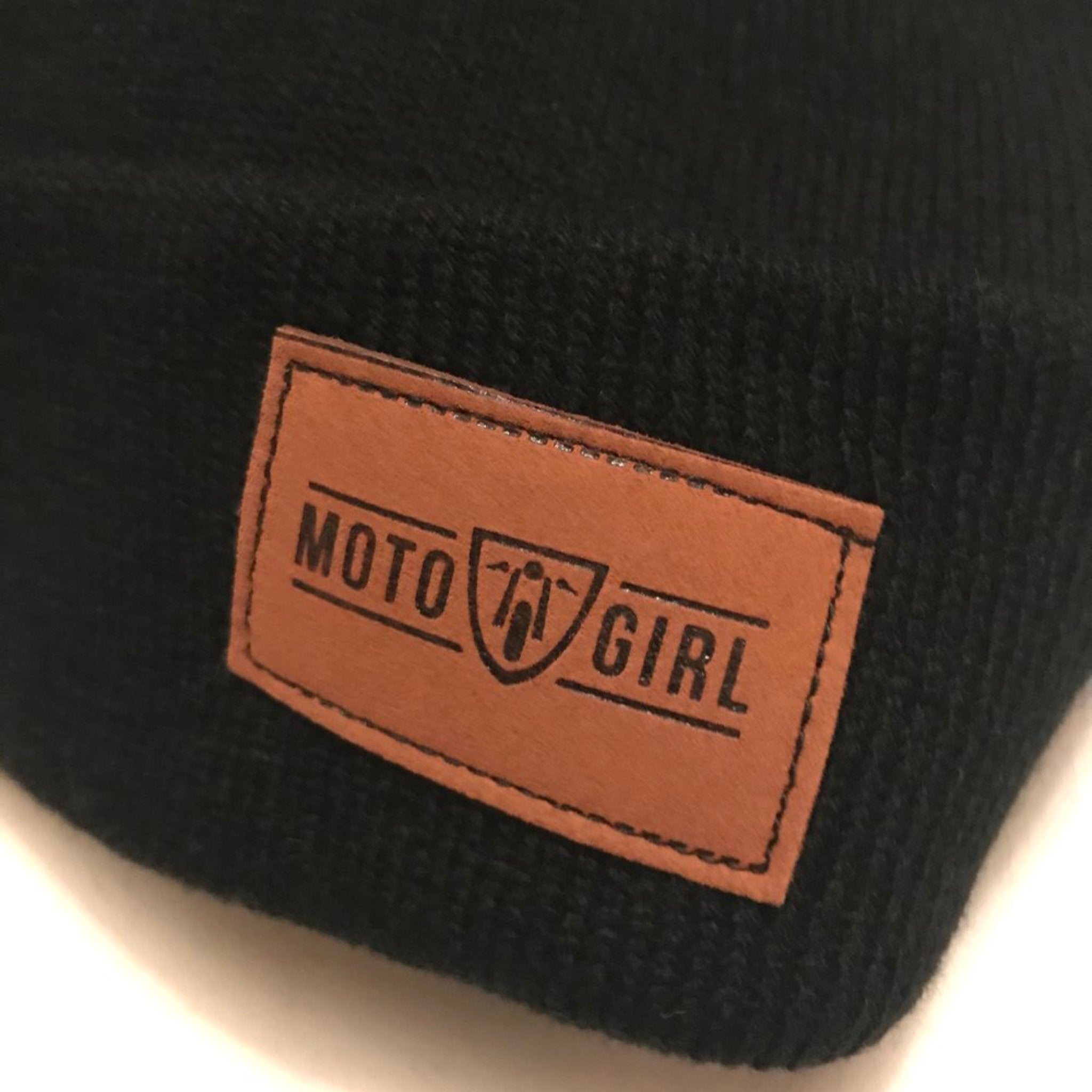 Close-up of the brown faux leather tag on the black beanie from MotoGirl, featuring the MotoGirl logo.