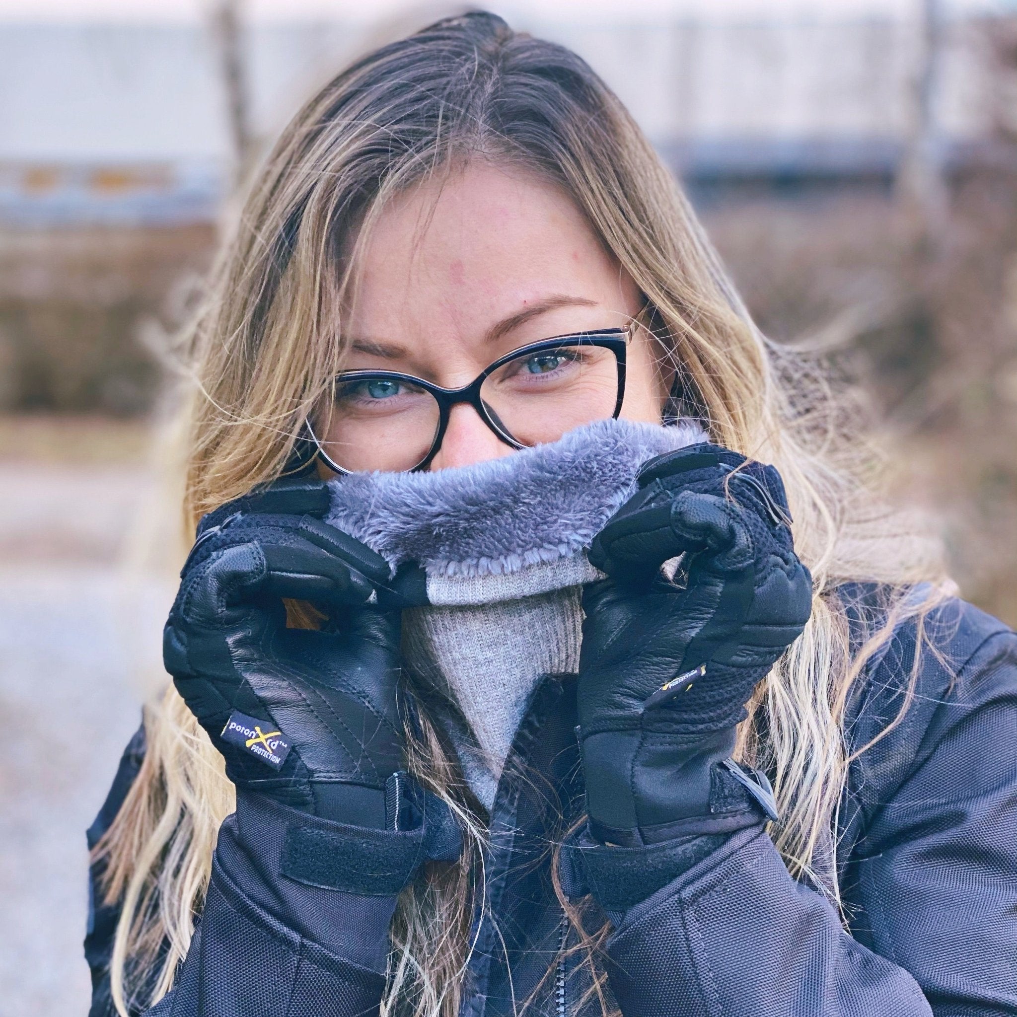 light haired woman in motorcycle gear, wearing a light gray neck warmer from Moto Girl outside, showing the inner lining of the neck warmer.