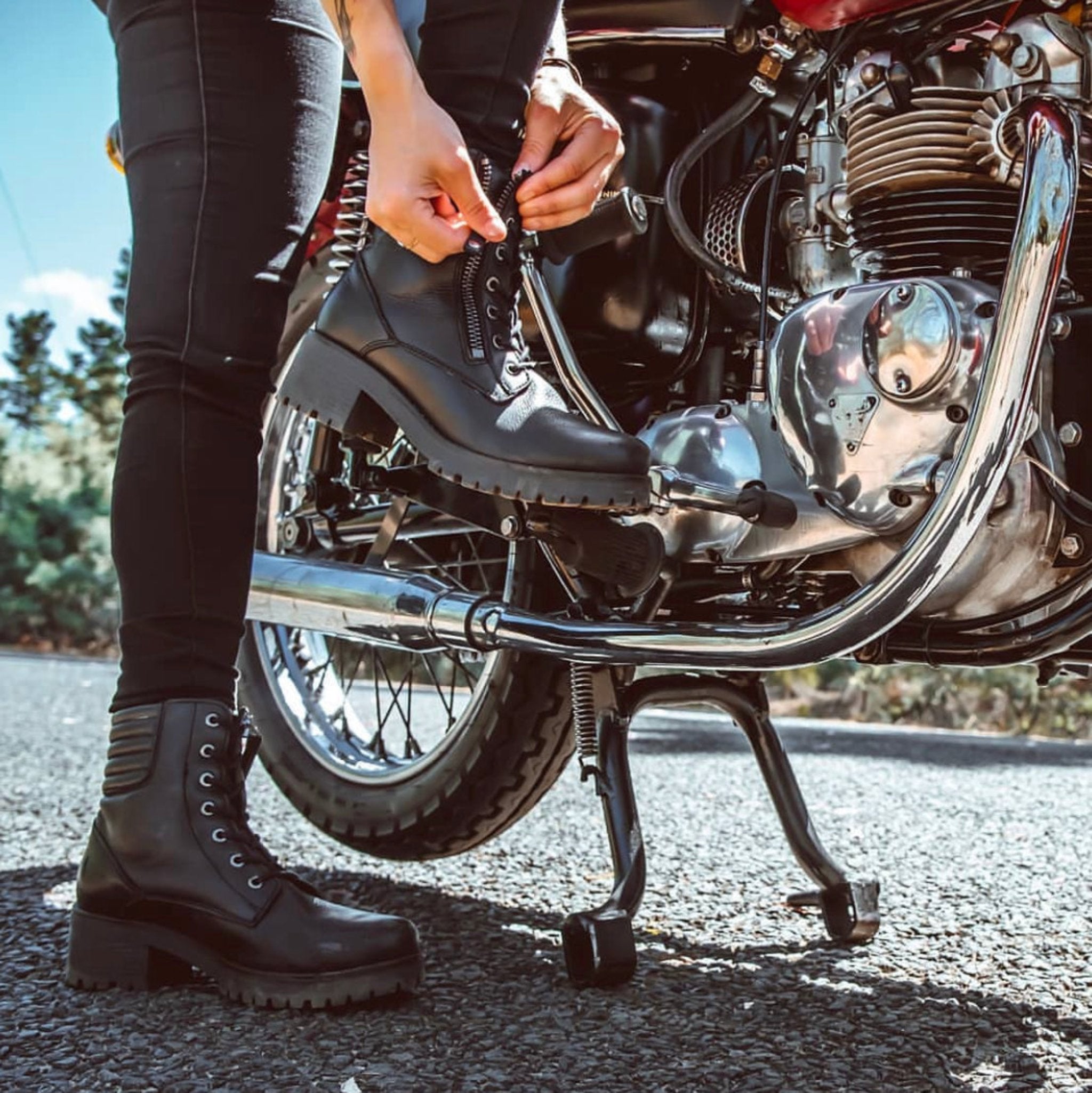 a woman standing by her motorcycle and zipping her black leather boot 