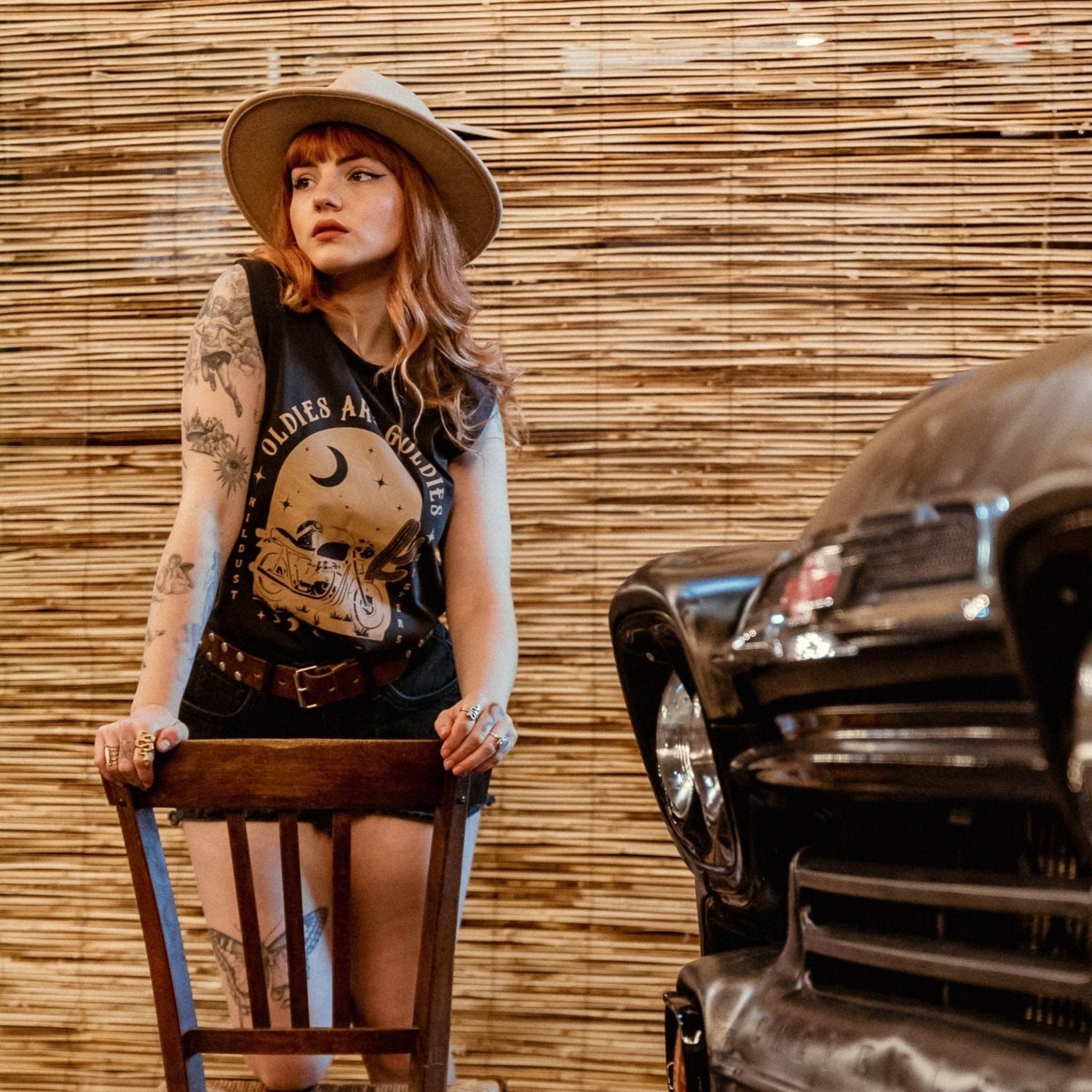 a young woman with a hat by a retro car wearing off-black tank top with brown motorcycle motive and oldies but goldies text