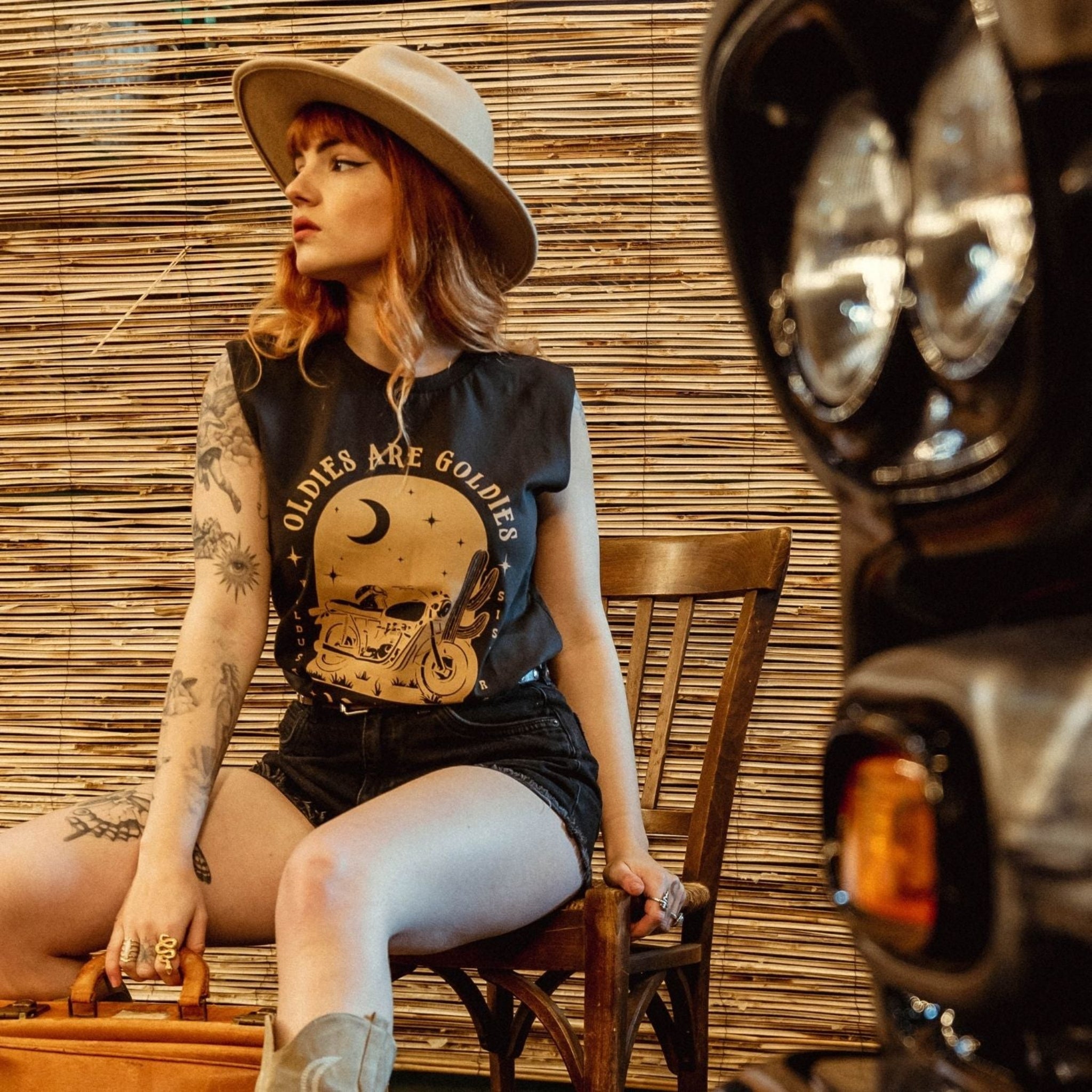 a young woman with a hat siting on a chair and wearing off-black tank top with brown motorcycle motive and oldies but goldies text