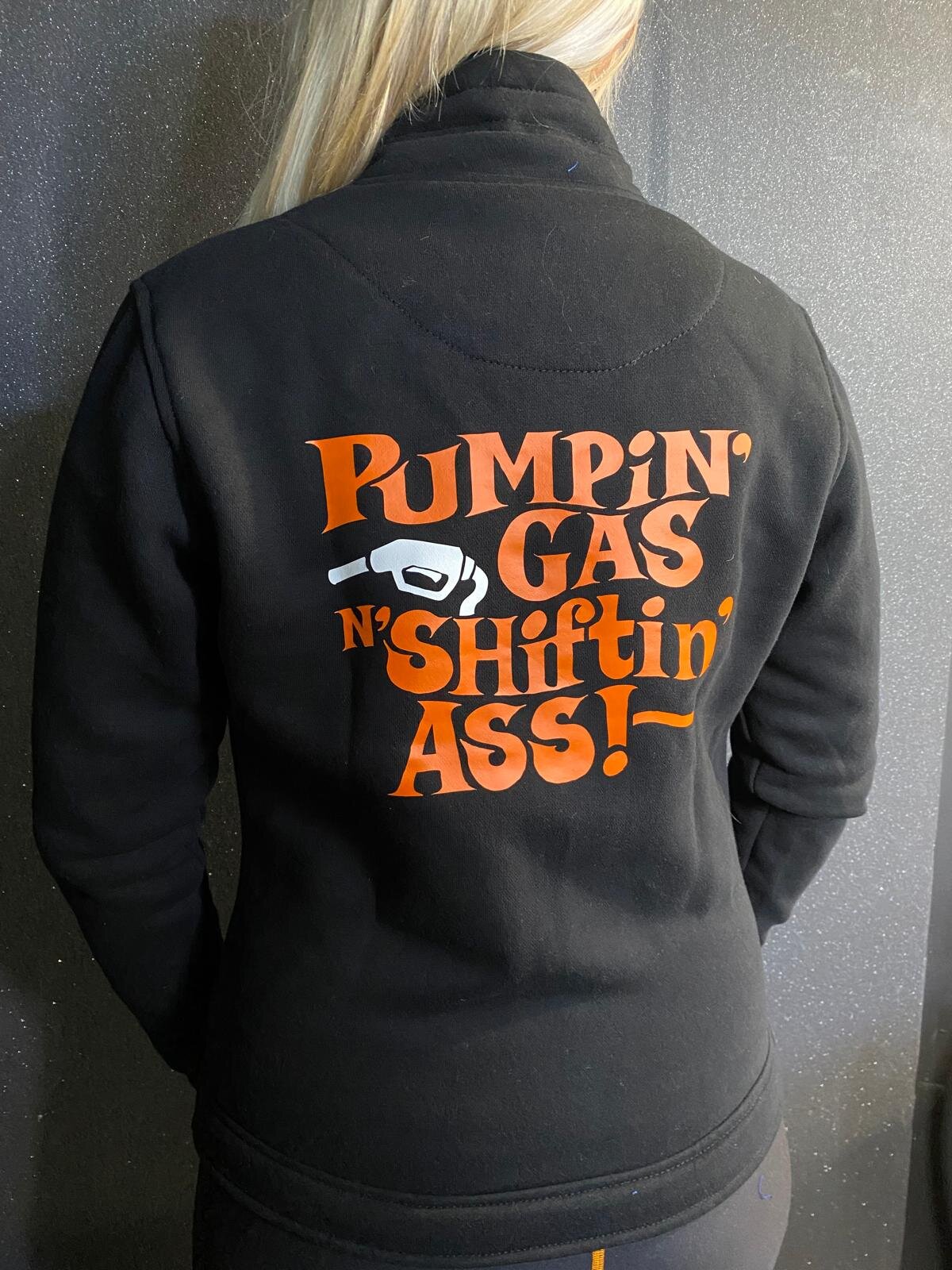 A woman wearing black lady sweatshirt with orange "pumping gas shifting ass"motive on the back