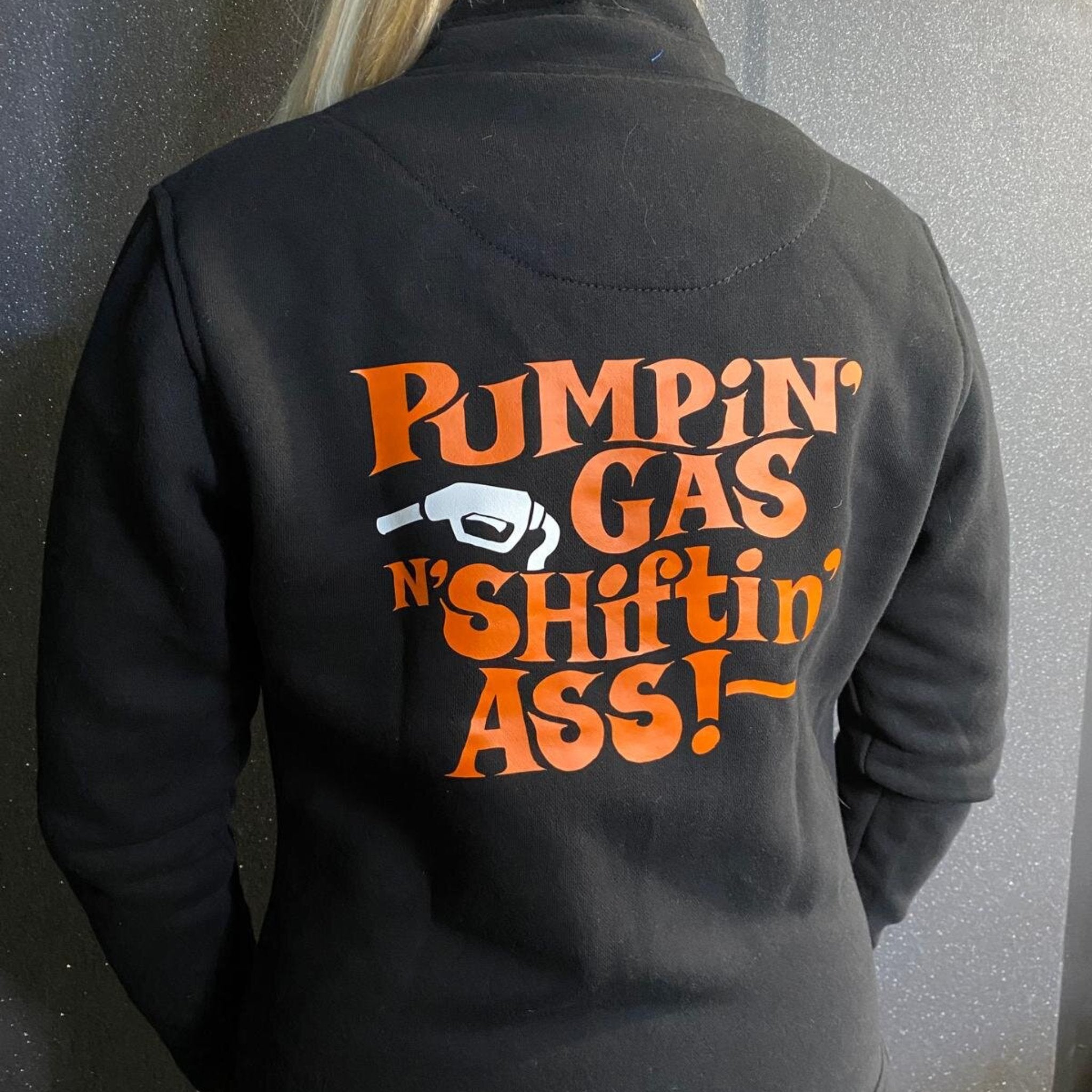 A woman wearing black lady sweatshirt with orange "pumping gas shifting ass"motive on the back