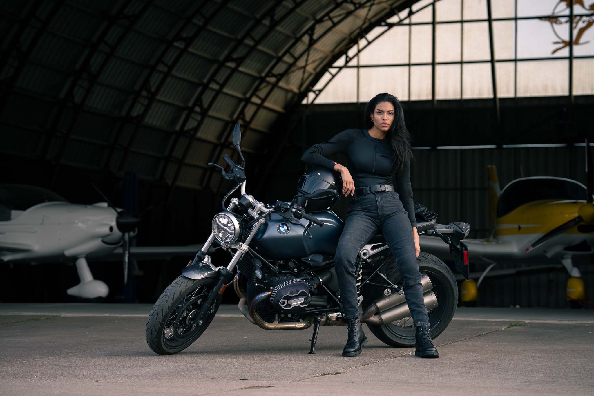 A woman in black outfit sitting on a motorcycle in a hangar with vehicles in the background