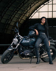 A woman in black outfit sitting on a motorcycle in a hangar with vehicles in the background