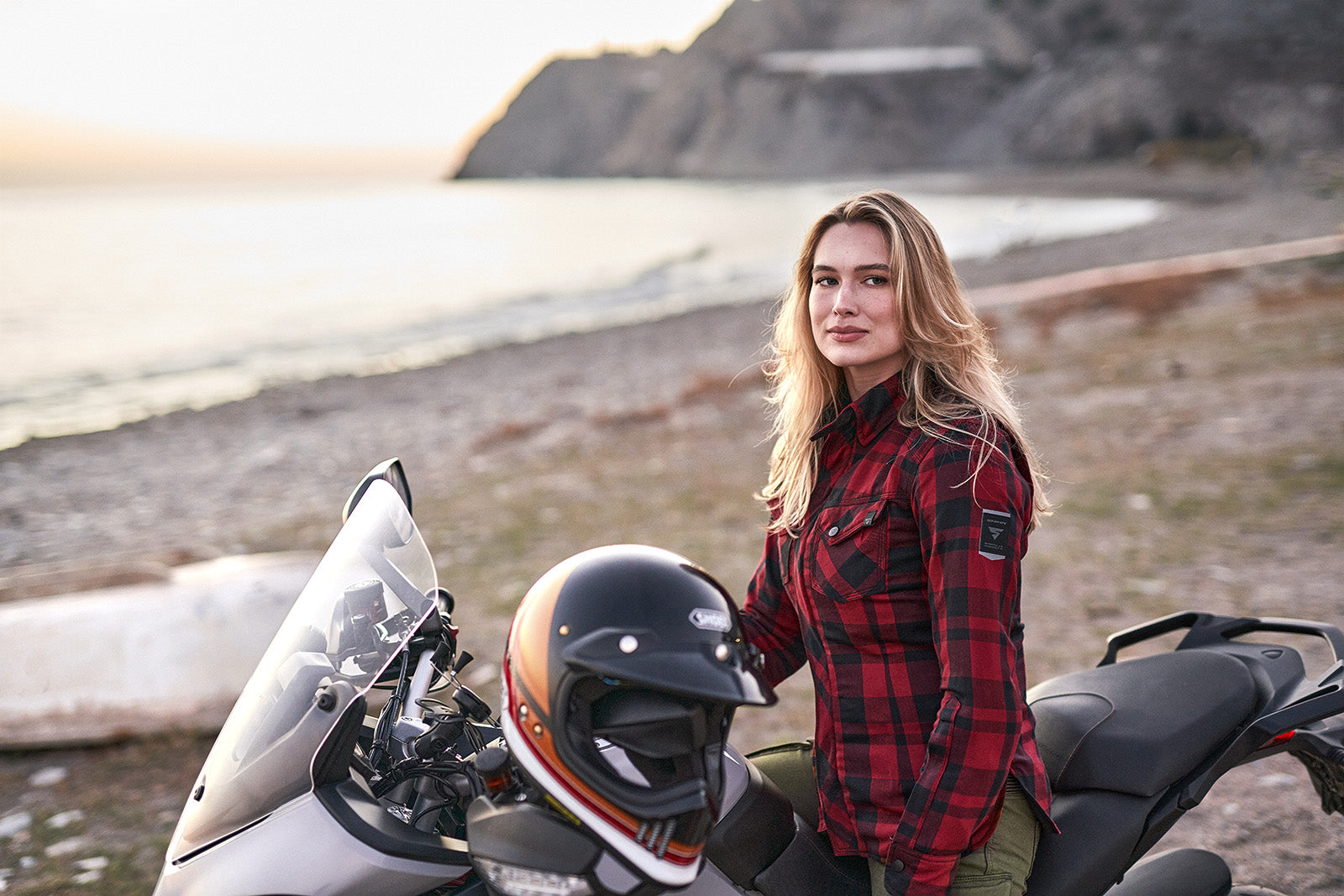 Woman in a plaid RED motorcycle shima jacket standing next to a motorcycle with a helmet on the seat, outdoors.