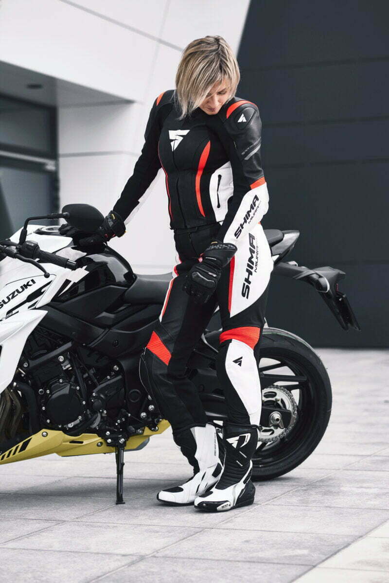 A woman standing next to her motorcycle wearing SHIMA MOTORCYCLE LEATHER suit IN BLACK FLUO, WHITE AND RED FLUO