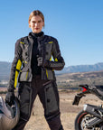 A young woman standing by her motorcycle wearing shima motorcycle touring jacket for women in black/ fluo