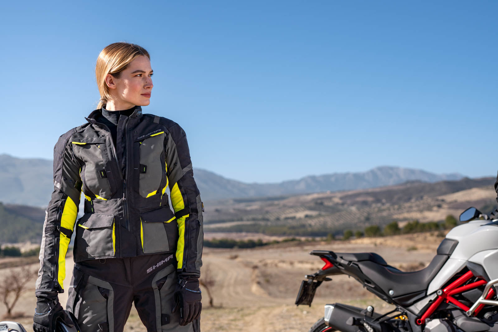 A woman standing by her mc wearing shima motorcycle touring jacket for women in black/ fluo