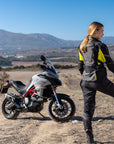 A woman standing by her motorcycle wearing Black SHIMA touring motorcycle pants for women