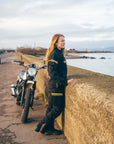 Person in motorcycle gear standing next to a motorcycle by a coastal road.