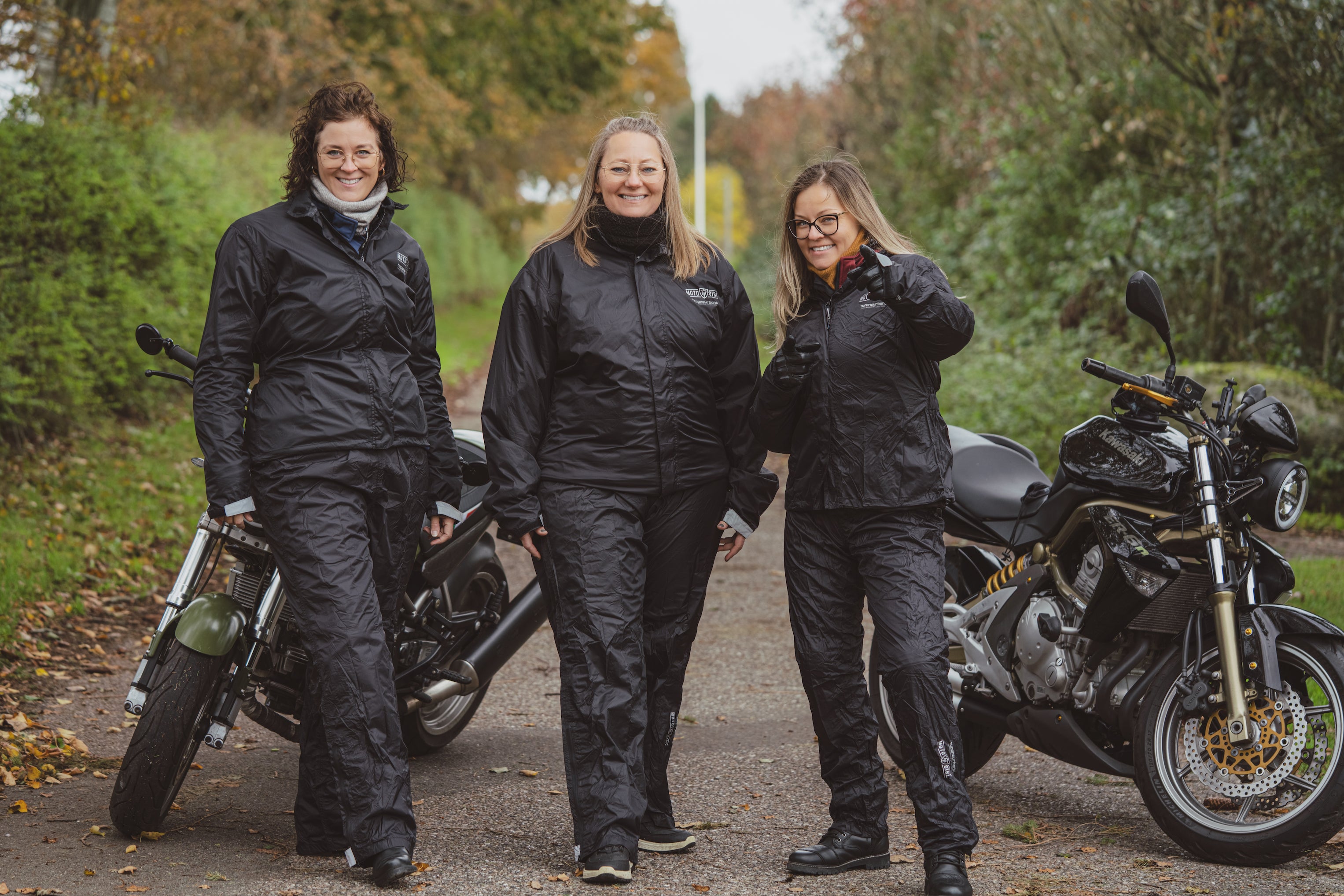 Three women standing by their motorcycles wearing black rain gear