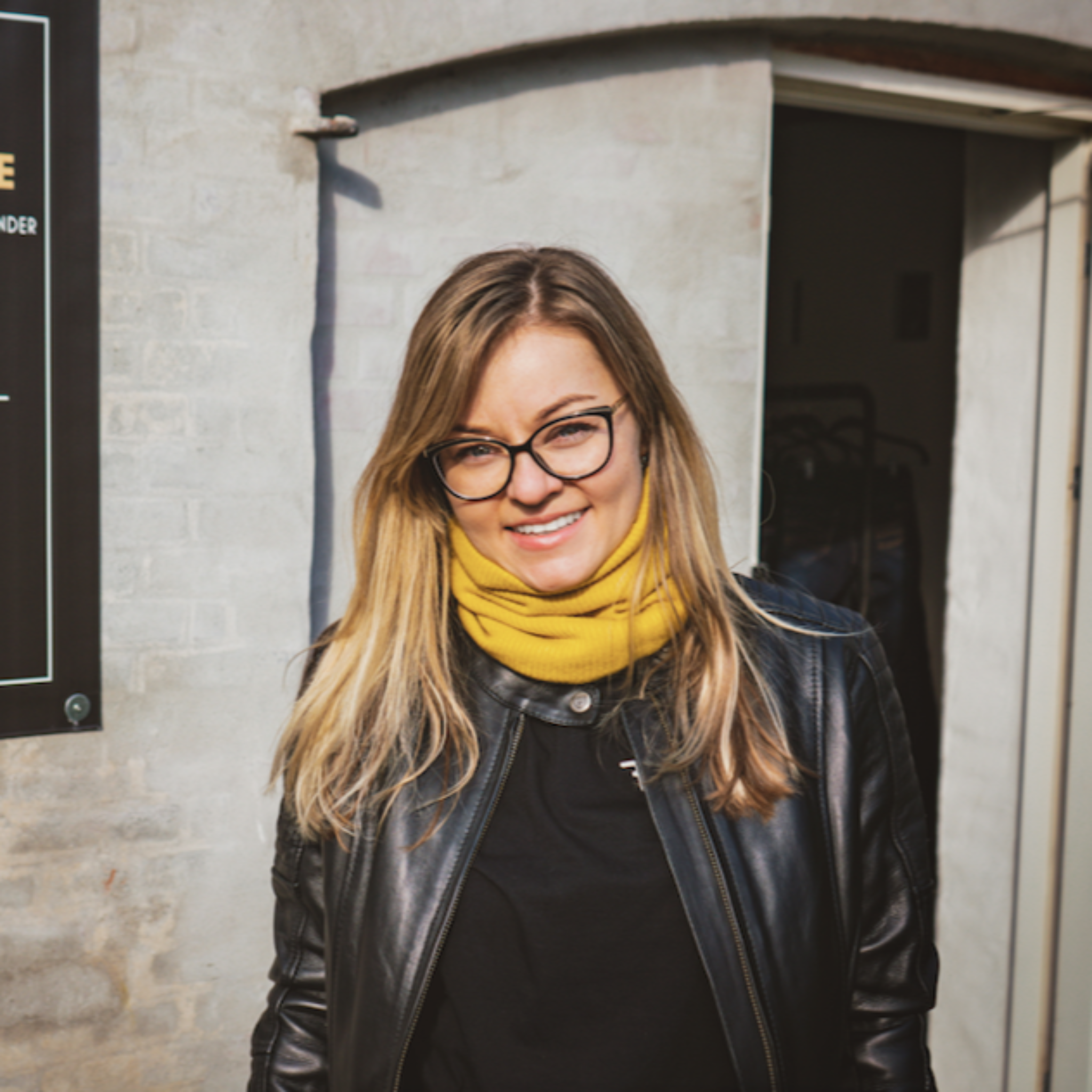 Smiling woman standing outside, wearing a black Valerie jacket and a yellow knitted neck warmer from Moto Girl.