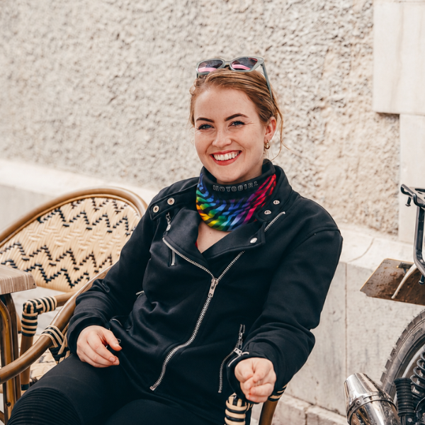 A smiling woman sits in a woven chair outdoors, wearing a black moto-style jacket, black pants, and a bright rainbow-striped neck gaiter. She has sunglasses on her head and a bicycle wheel visible on the right side of the frame.